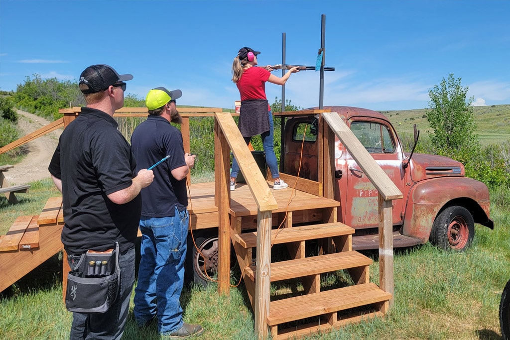 Woman shooting a shotgun at a stand with a man holding a controller and another holding a clipboard